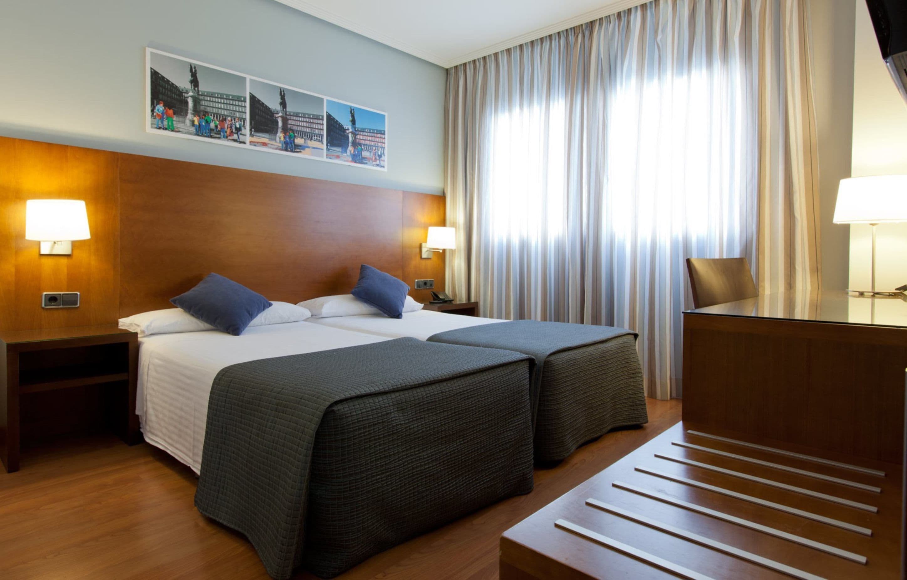 A hotel room with two beds, a desk, and a chair. There are two wall-mounted lamps and a large window with curtains.