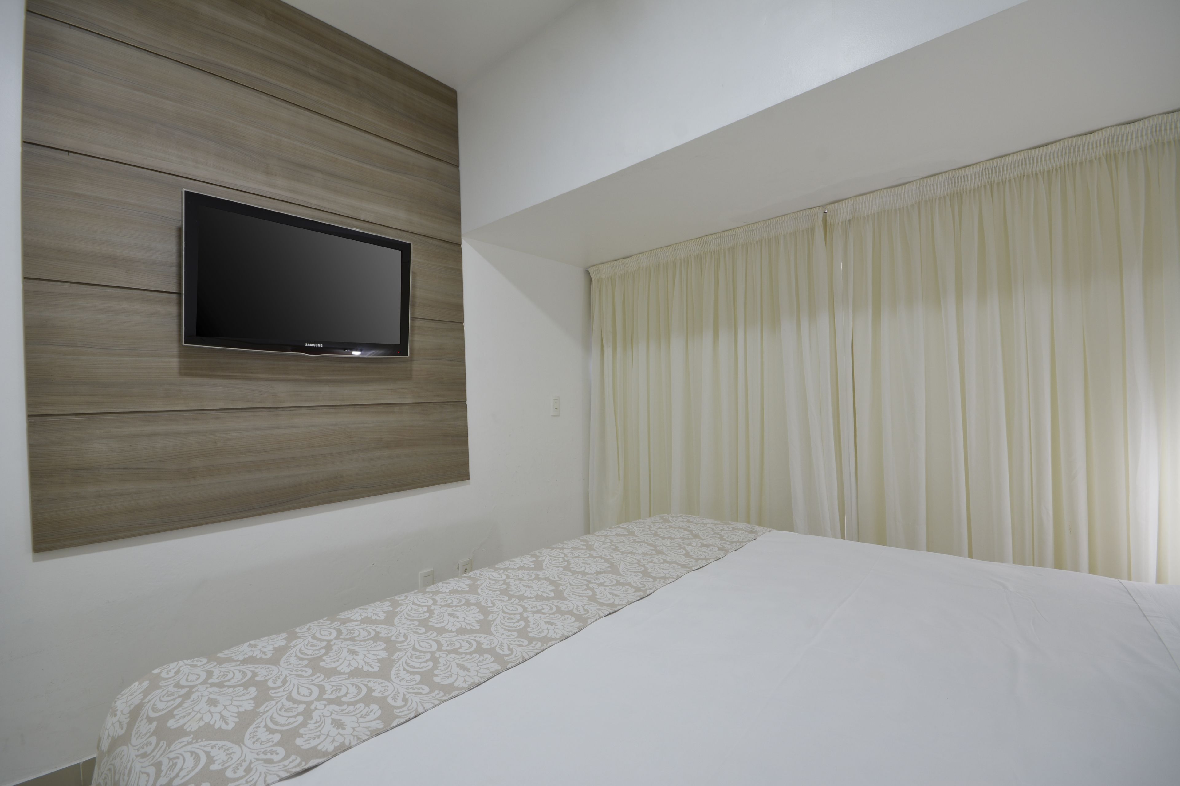 Habitación de hotel con cama, televisor montado en una pared de paneles de madera y ventana con cortinas.