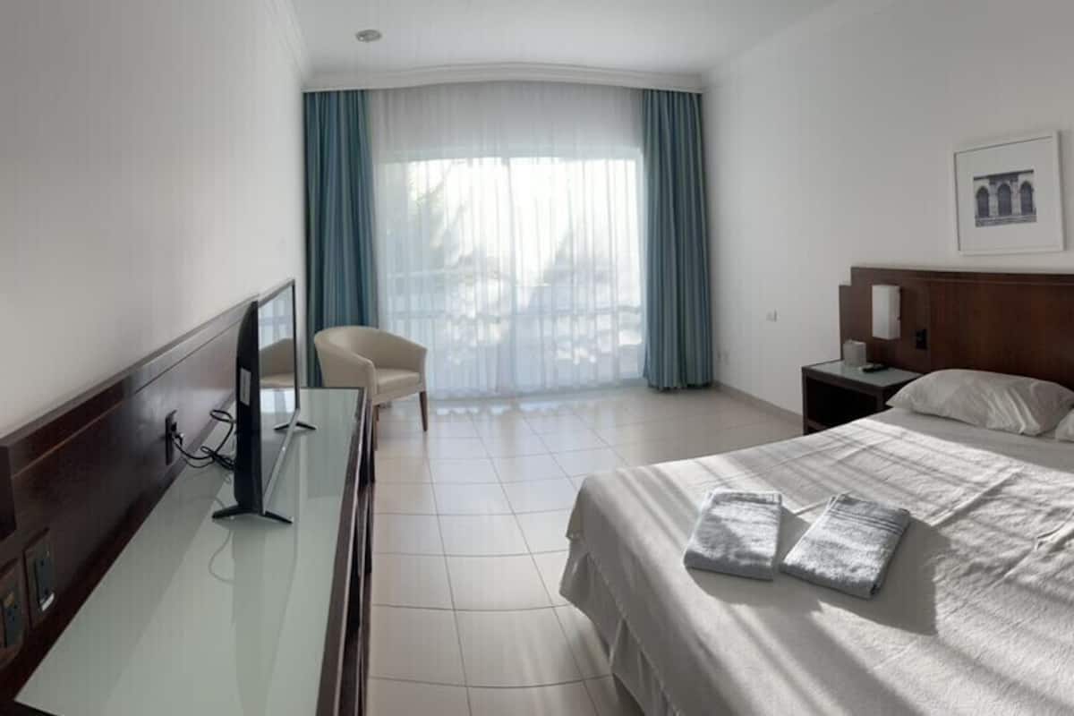 Habitación de hotel con cama, mesitas de noche, un escritorio con computadora, una silla y un ventanal con cortinas.