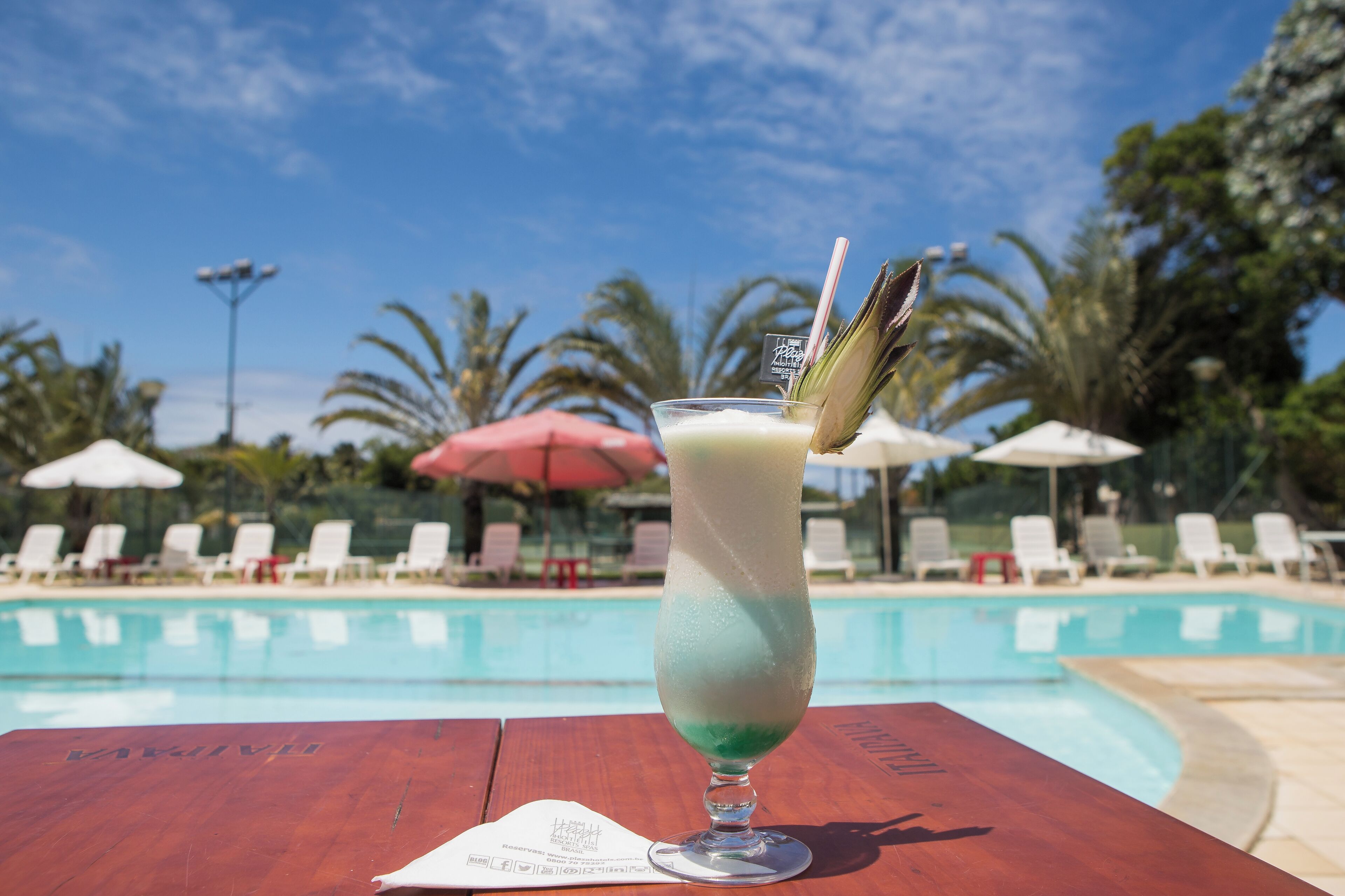 A poolside setting with a drink in a tall glass, a wooden table, and lounge chairs with umbrellas.