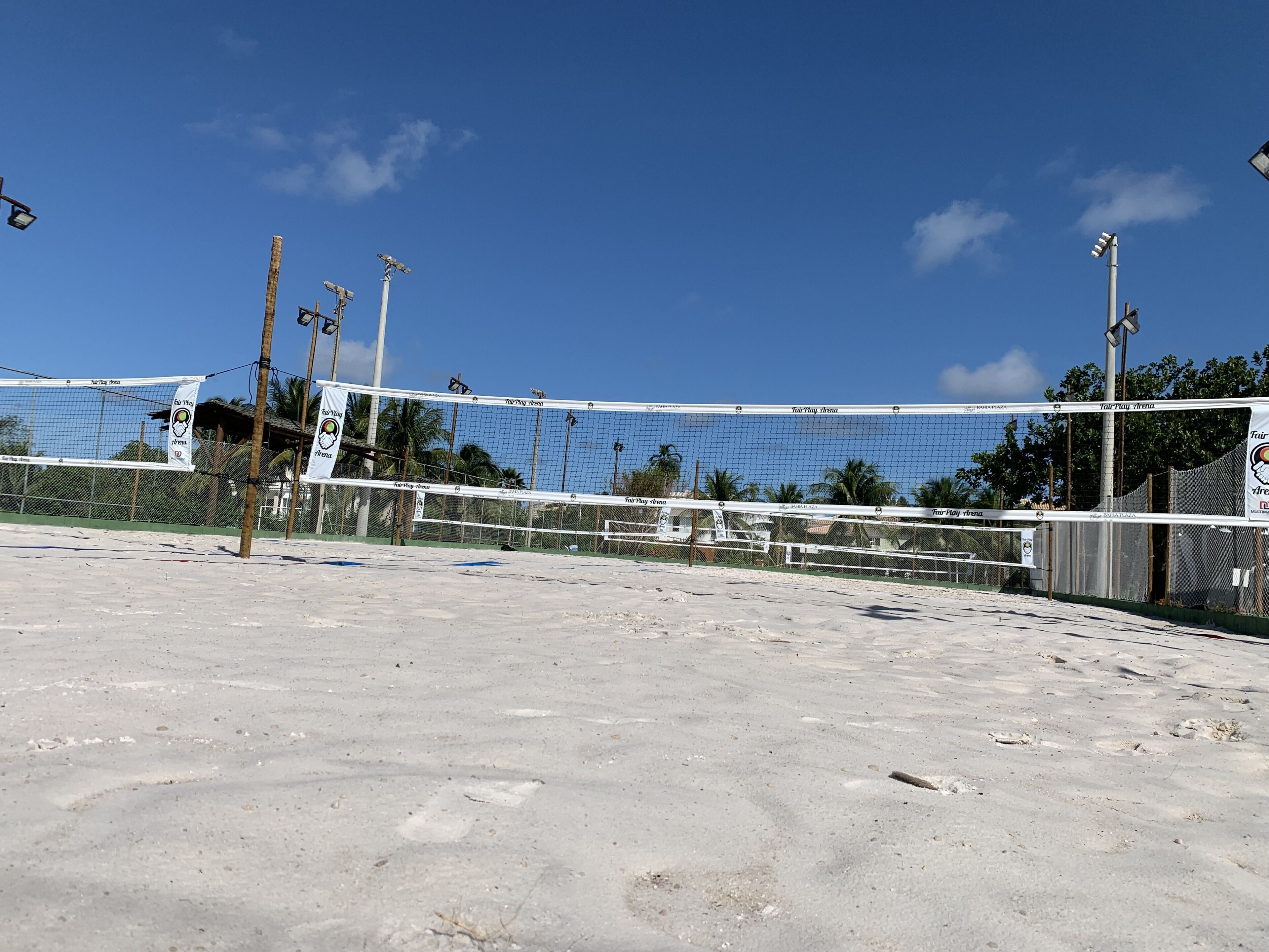 A sandy beach volleyball court with a net, surrounded by palm trees and a clear blue sky.