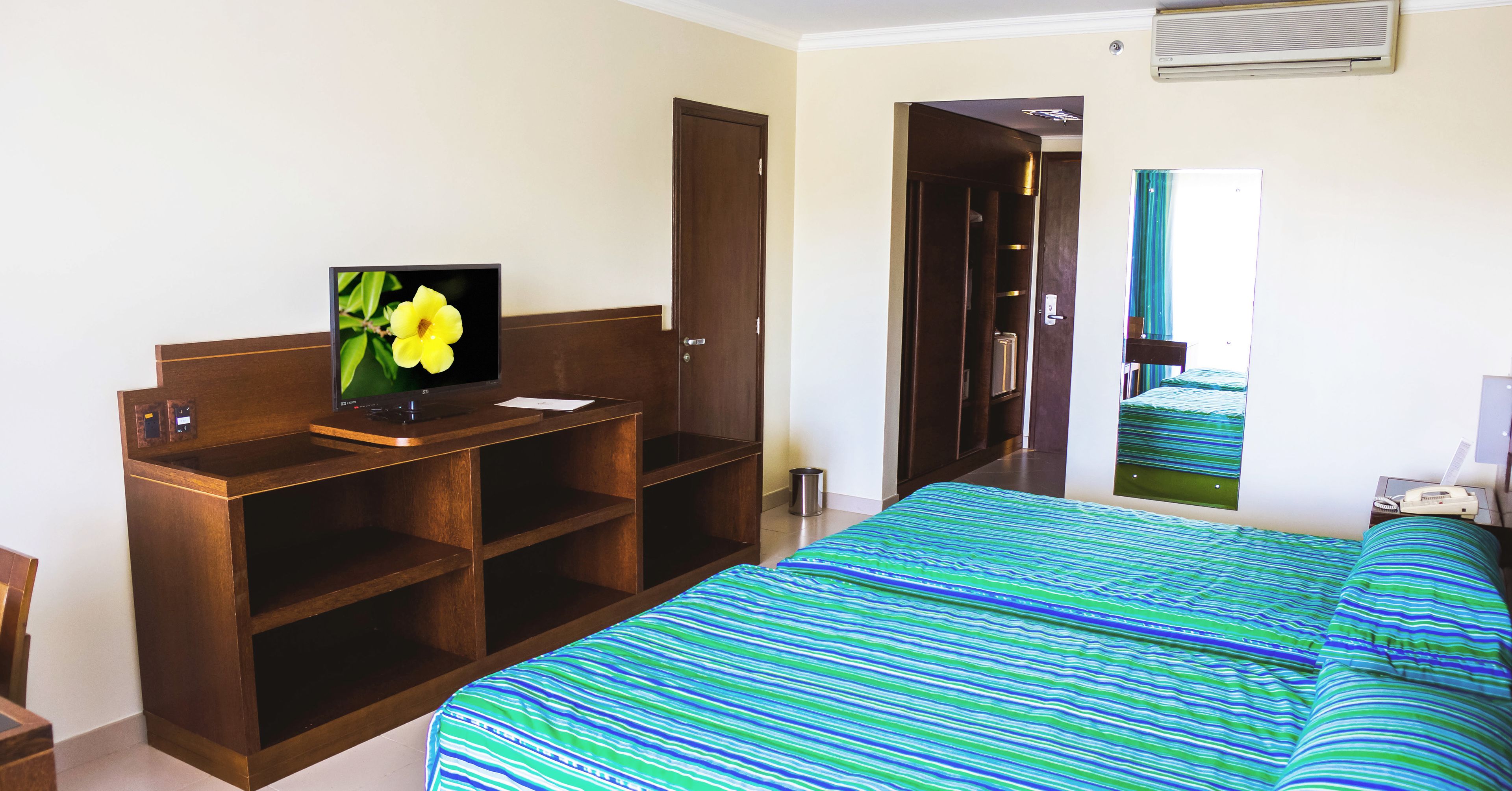 Une chambre d’hôtel avec un lit, un téléviseur posé sur un meuble en bois, un miroir et une porte donnant sur une autre pièce.