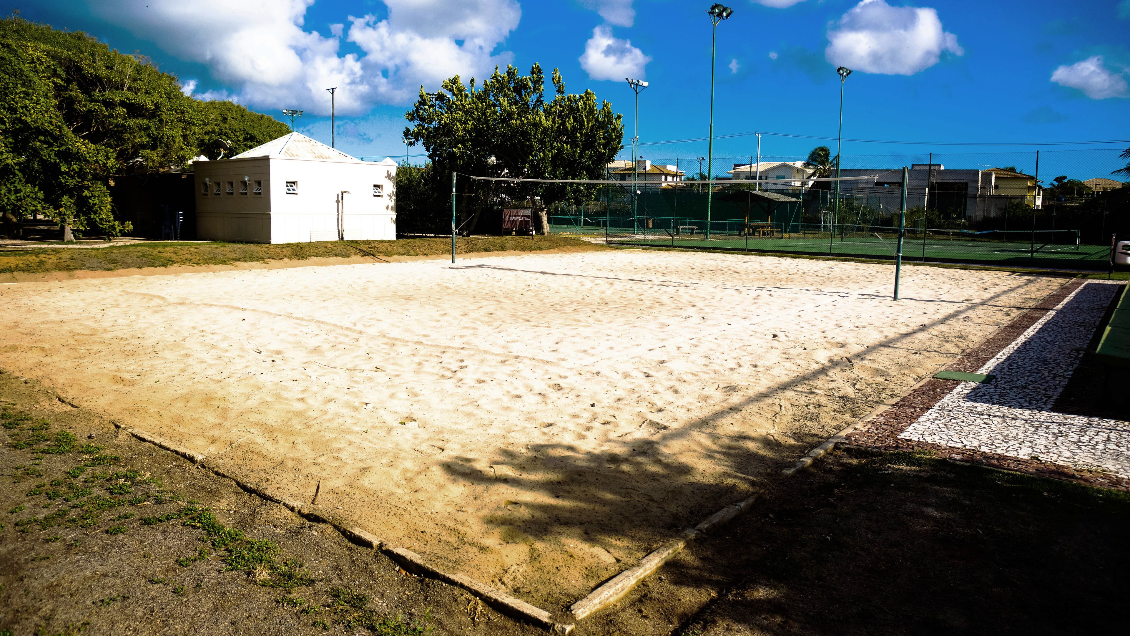 A sandy sports court with a net, surrounded by a fence, and a building in the background.