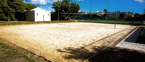 A sandy sports court with a net, surrounded by a fence, and a building in the background.