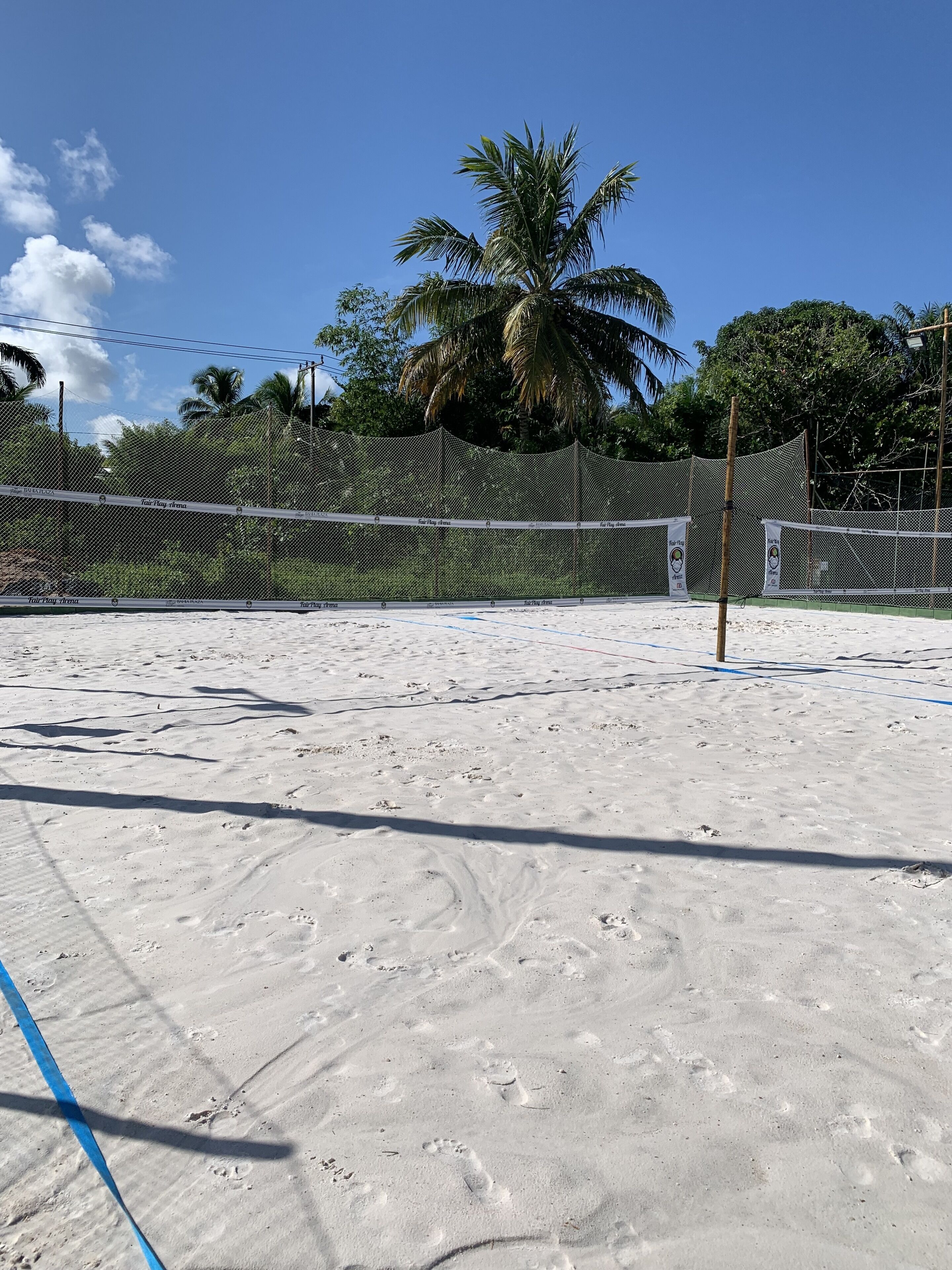 Een zandstrand met een volleybalveld, voetsporen, een volleybalnet en palmbomen op de achtergrond.