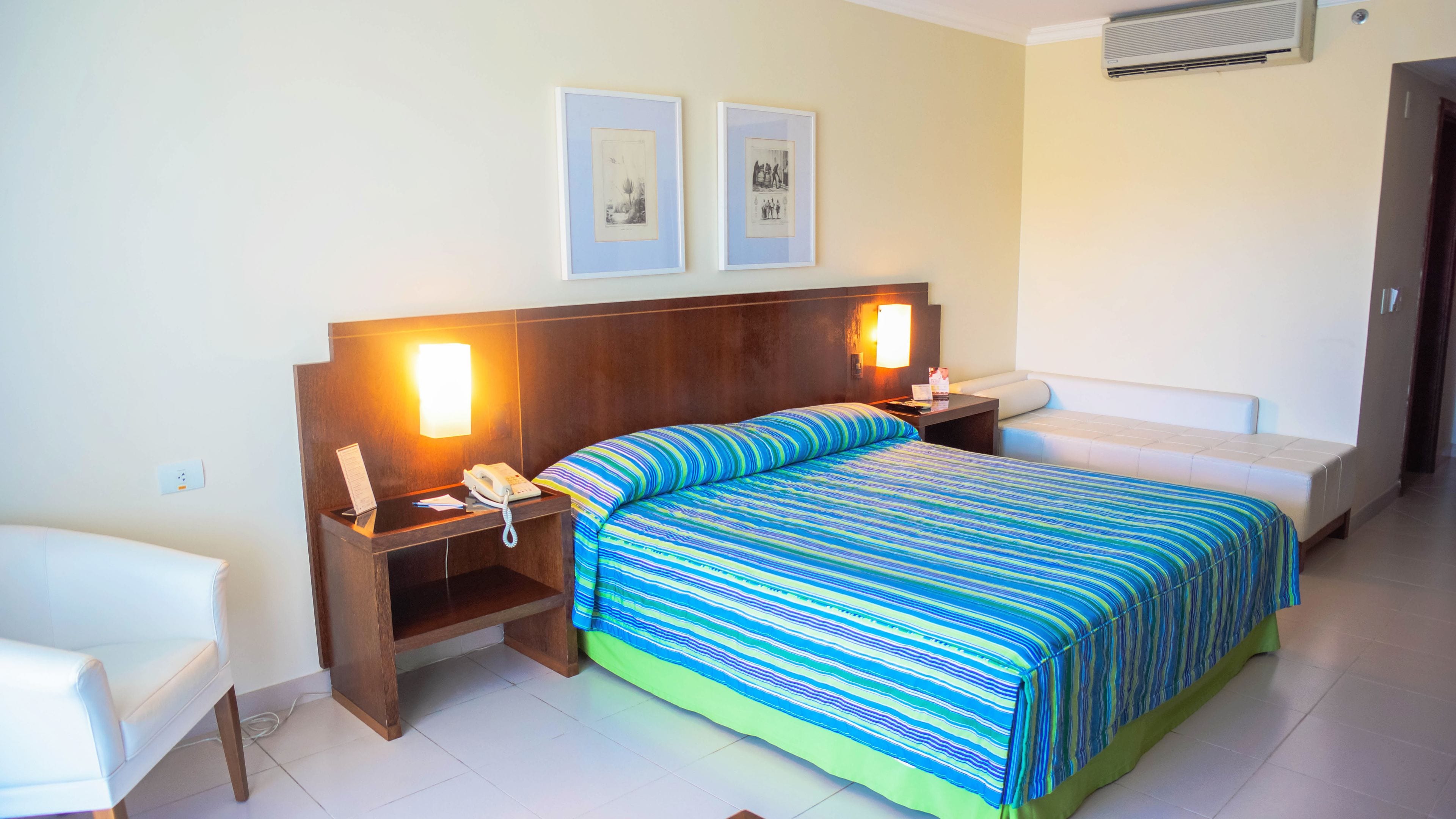 Una habitación de hotel con una cama grande, dos mesitas de noche con lámparas, una silla y dos cuadros en la pared.