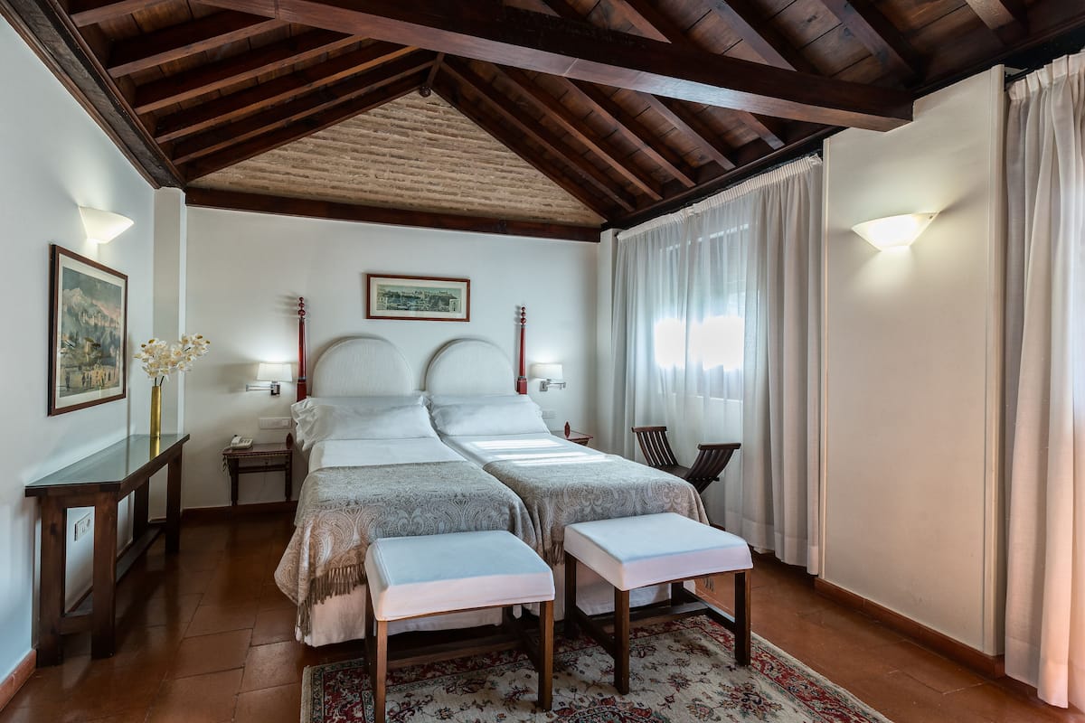 A double room with two beds, a wooden ceiling, a nightstand with a lamp, a framed picture on the wall, and a window with curtains.