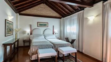 Habitación doble con dos camas, techo de madera, mesita de noche con lámpara, cuadro enmarcado en la pared y ventana con cortinas.