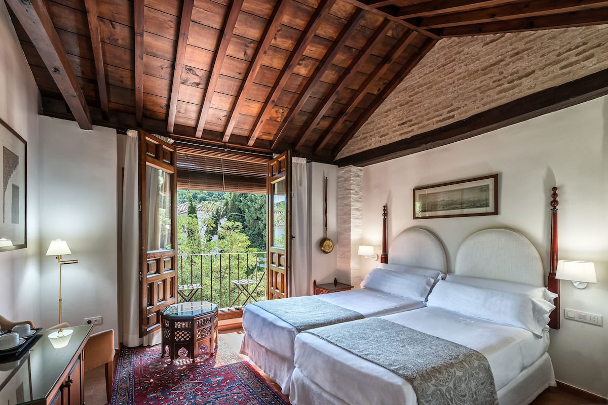 A room with two beds, a wooden ceiling, a balcony with a view of greenery, and a patterned rug.