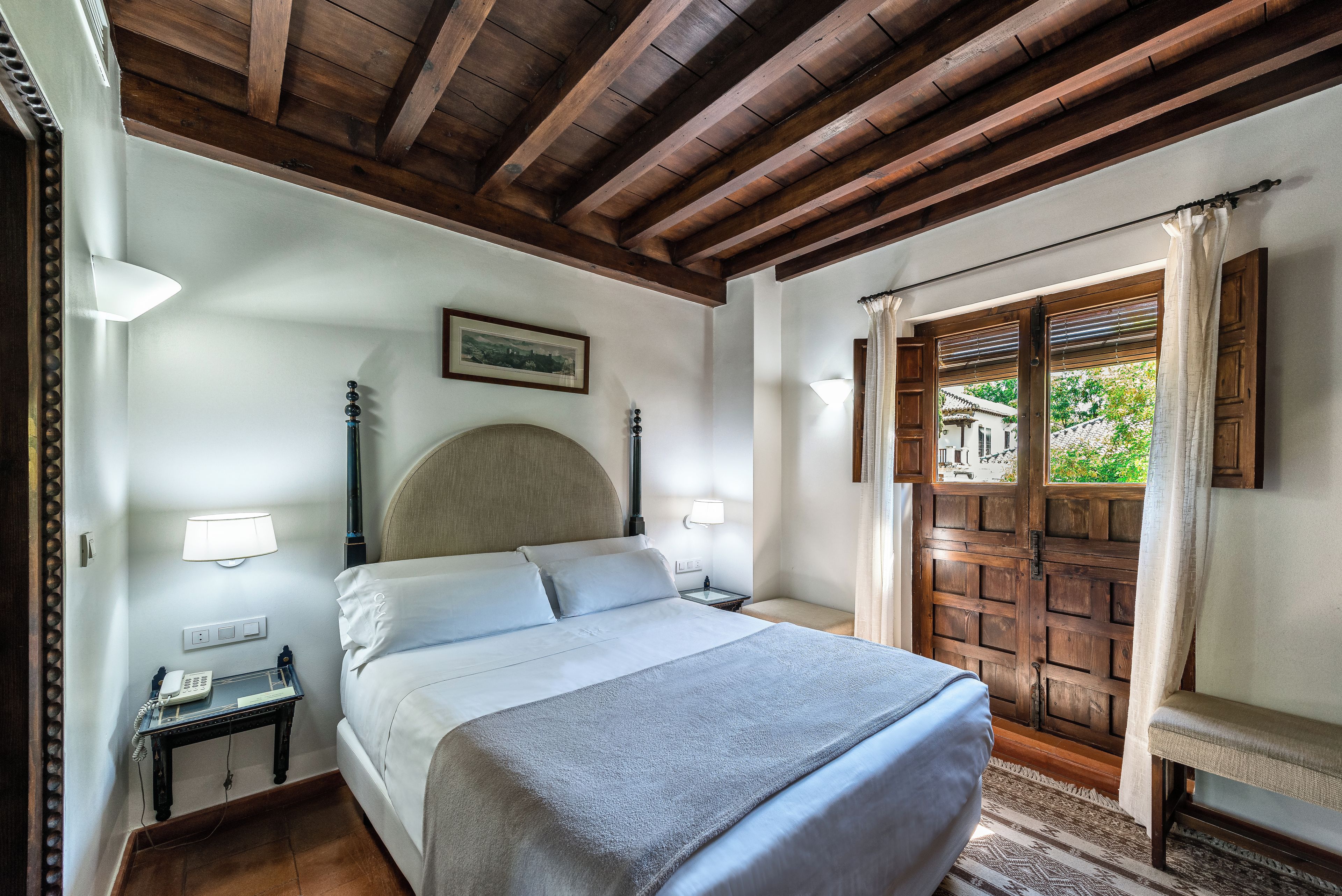 A hotel room with a bed, bedside lamps, a nightstand, a framed picture on the wall, and a window with curtains.