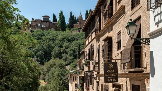 A historic building with a sign for Casa Morisca Hotel, featuring traditional architecture and a view of a hill with trees and a church. - Casa Morisca Hotel Boutique (Granada)