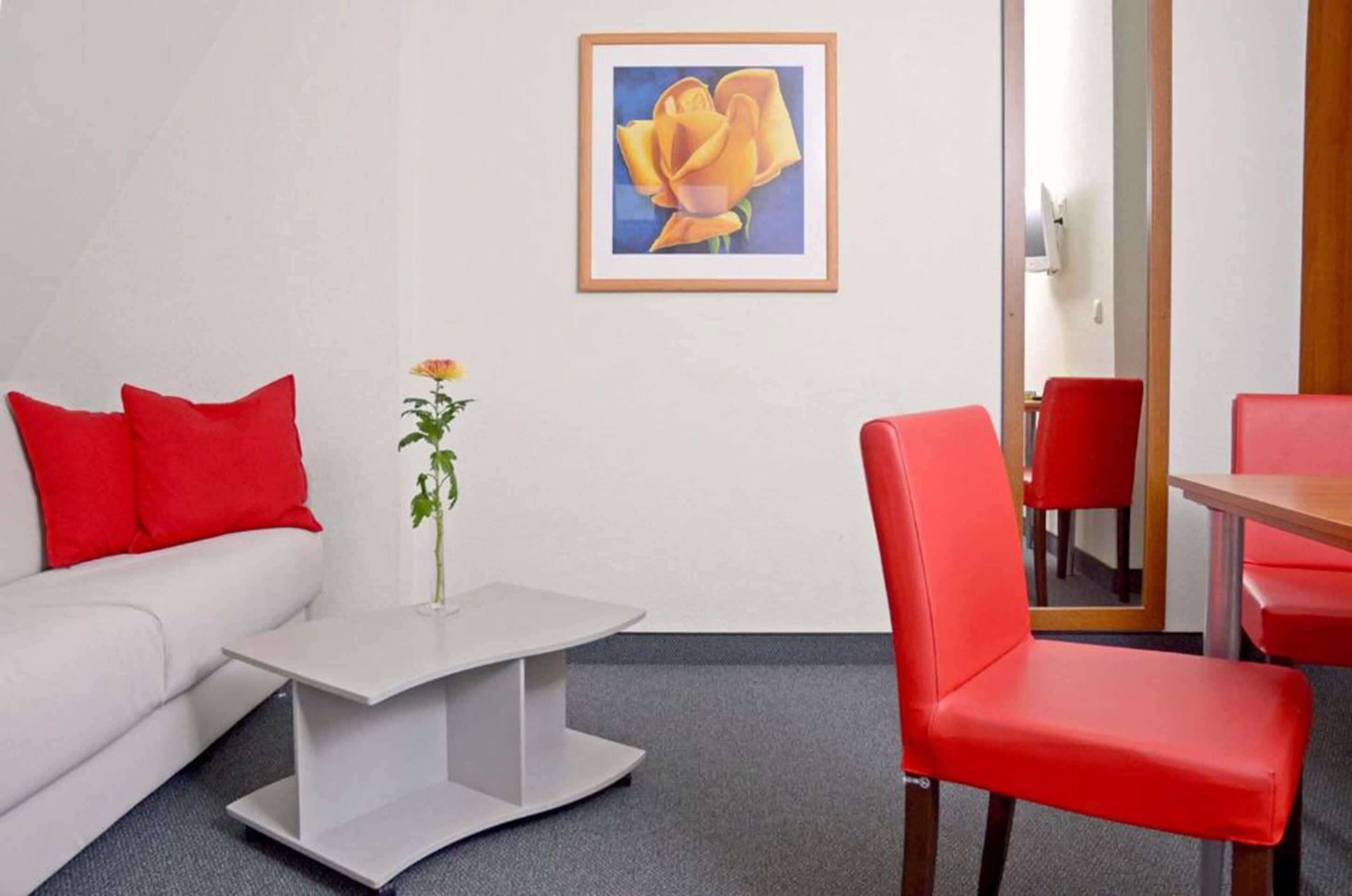 Une chambre d’hôtel avec un canapé blanc, des coussins rouges, une petite table blanche ornée d’un vase de fleurs, une chaise rouge et un tableau encadré représentant une fleur accroché au mur.