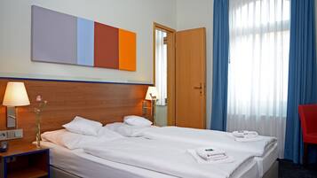 A hotel room with a bed, a nightstand with a lamp, a vase with flowers, a mirror, a door, and a window with curtains.