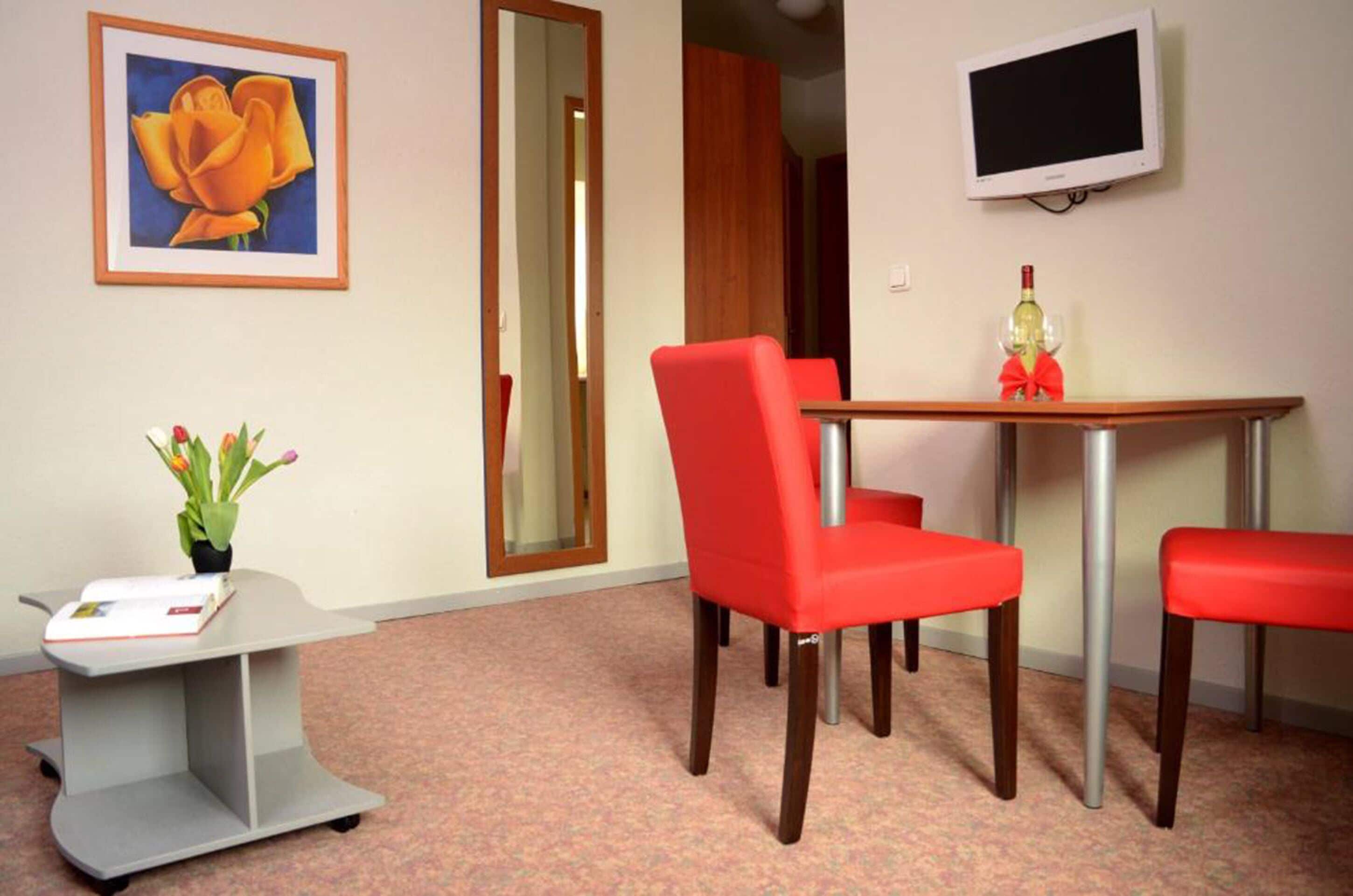 Una habitación con una mesa de madera, sillas rojas, un televisor pequeño, un cuadro enmarco y un escritorio con una planta.