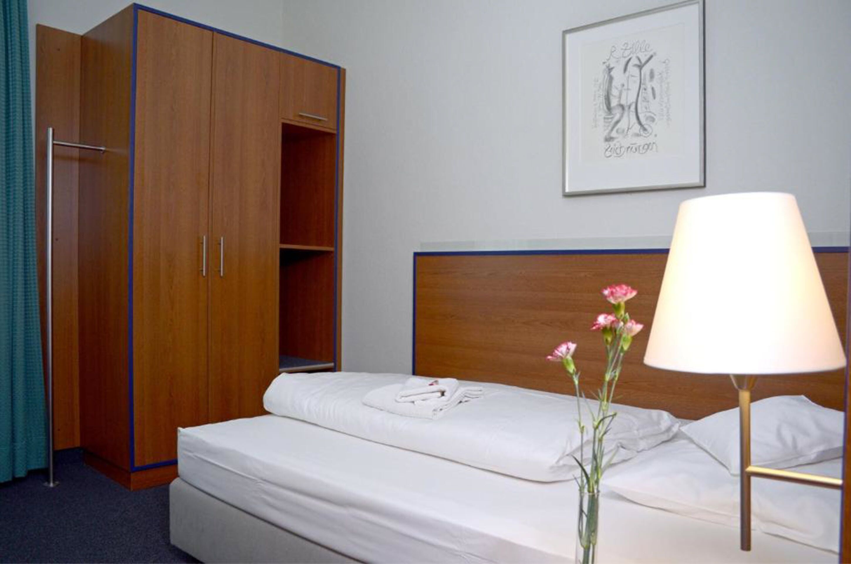 A hotel room with a bed, a wooden wardrobe, a framed artwork on the wall, and a bedside lamp.