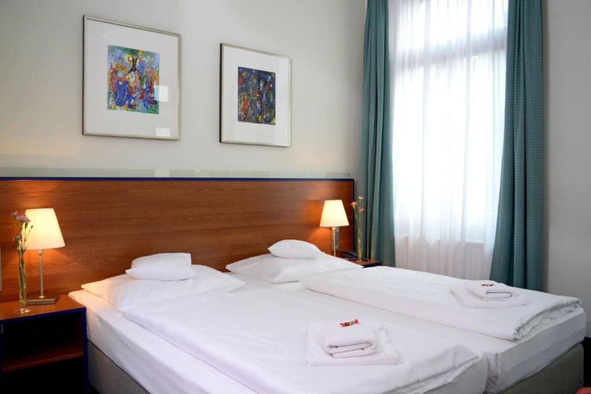 A hotel room with a large bed, two framed artworks on the wall, and a window with curtains.