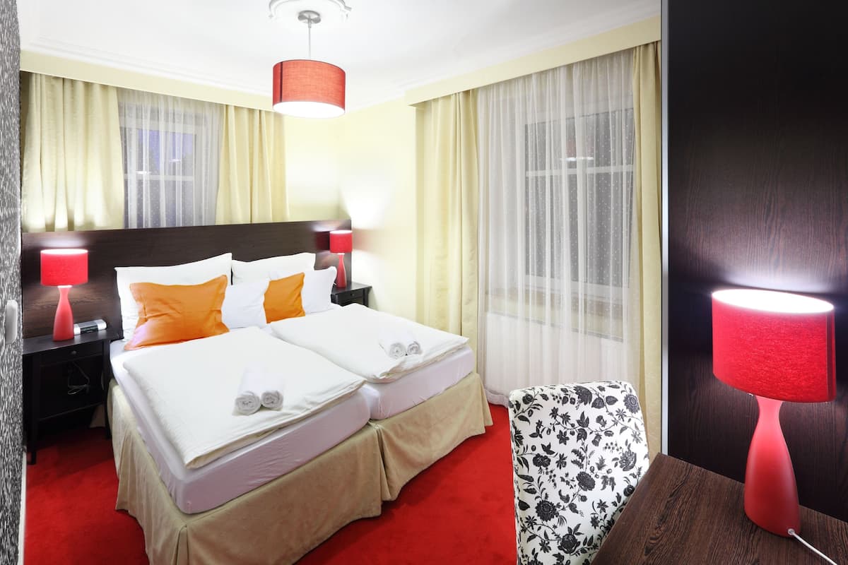 A hotel room with a double bed, two bedside tables, a red lamp, a floral chair, and a large window with sheer curtains.