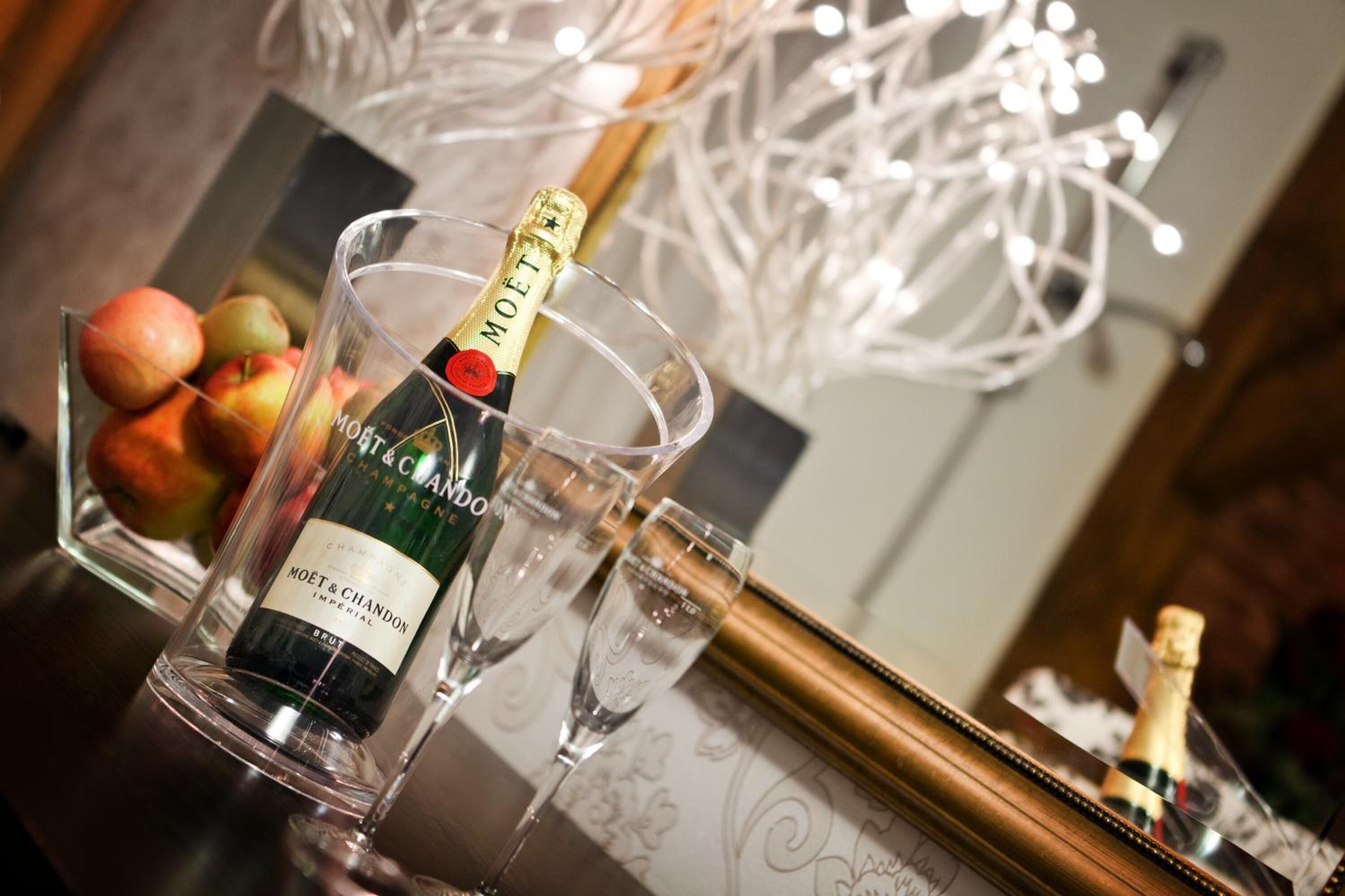 A bottle of Moët & Chandon champagne in a glass, with a bowl of fruit and a framed picture in the background.