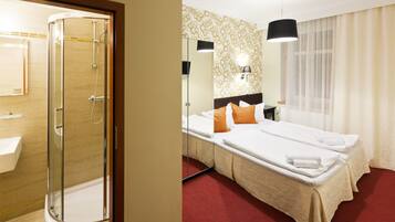 A hotel room with a glass-enclosed shower, a bed with white linens, an orange pillow, a bedside table, and a window with sheer curtains.