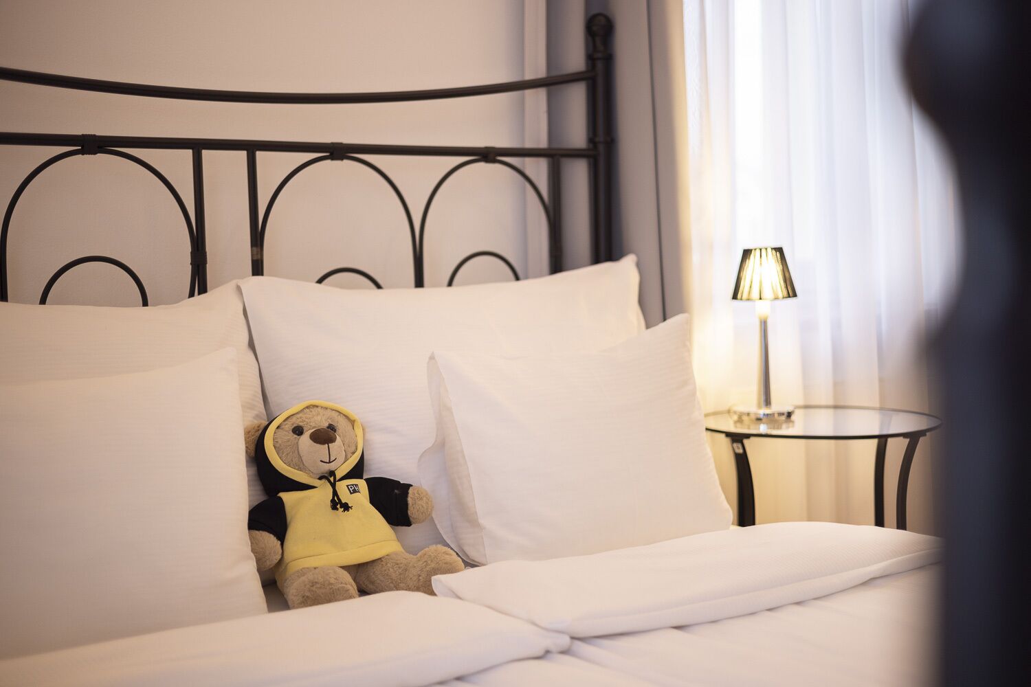 Een net opgemaakt bed met witte lakens, een teddybeer met een gele hoodie, en een bedlamp op een klein rond tafeltje.