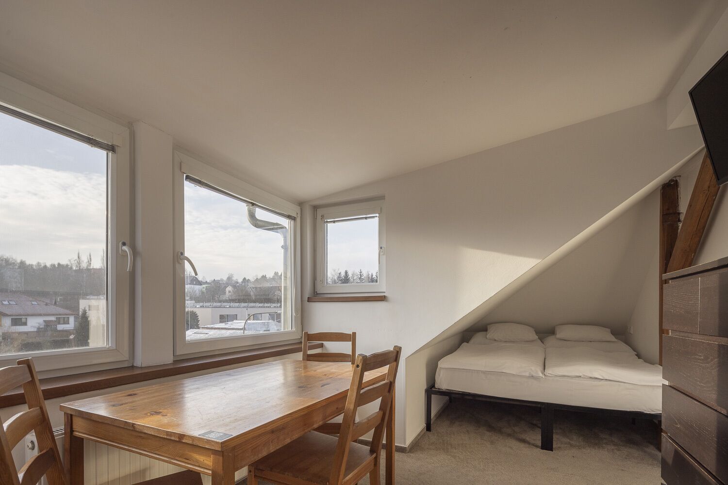 A small attic bedroom with a wooden table and chairs, a bunk bed, and large windows offering views of the neighborhood.