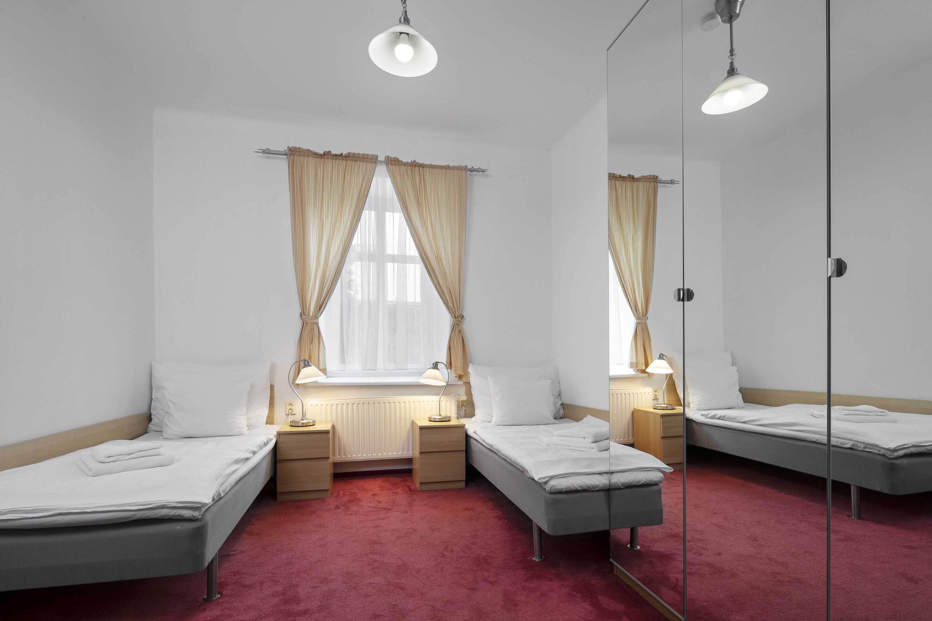 Een kamer met drie eenpersoonsbedden, een raam met gordijnen, nachtlampjes en een radiator.