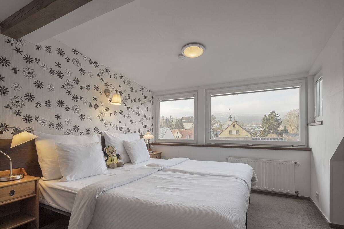 Un dormitorio con cama, mesita de noche, lámpara, un osito de peluche y ventana con vista a edificios.