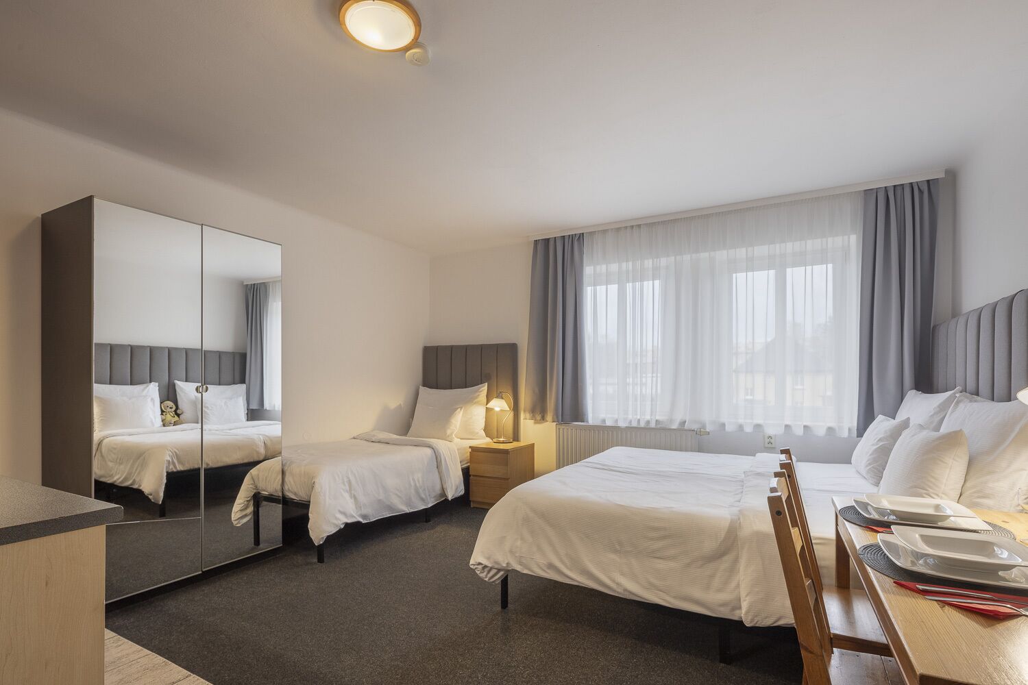 A hotel room with two beds, a large mirror, a bedside table with a lamp, a window with curtains, and a dining area with a table and chairs.