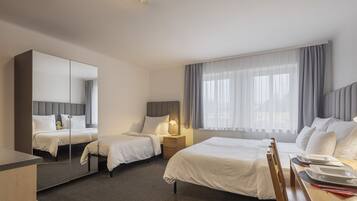 A hotel room with two beds, a large mirror, a bedside table with a lamp, a window with curtains, and a dining area with a table and chairs.
