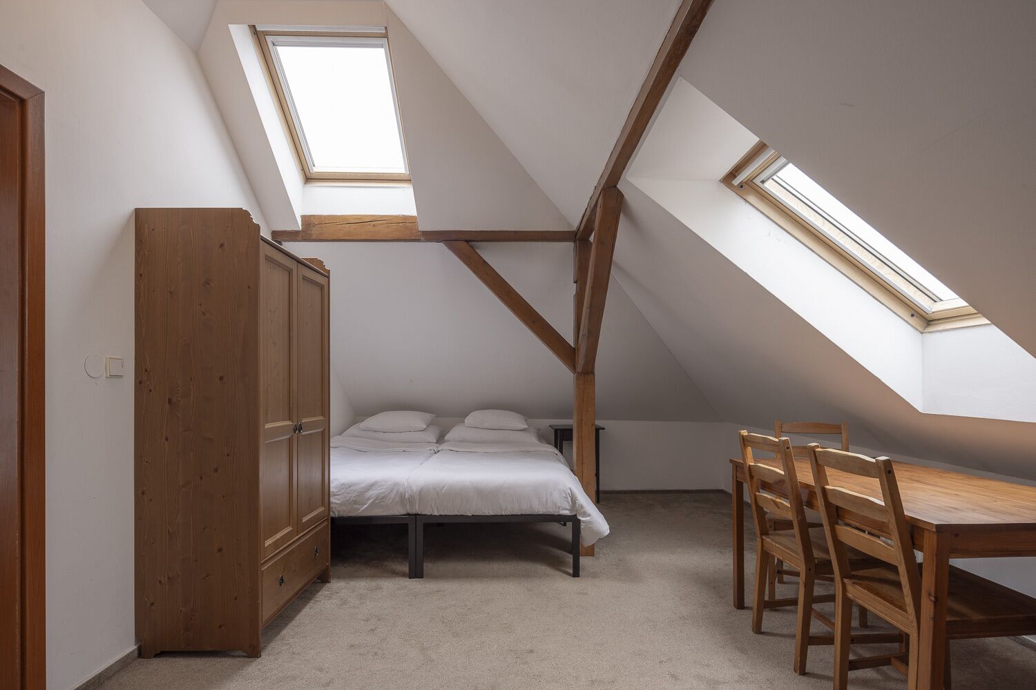 A small attic bedroom with a sloped ceiling, a bed, a wooden wardrobe, and a dining table with chairs.