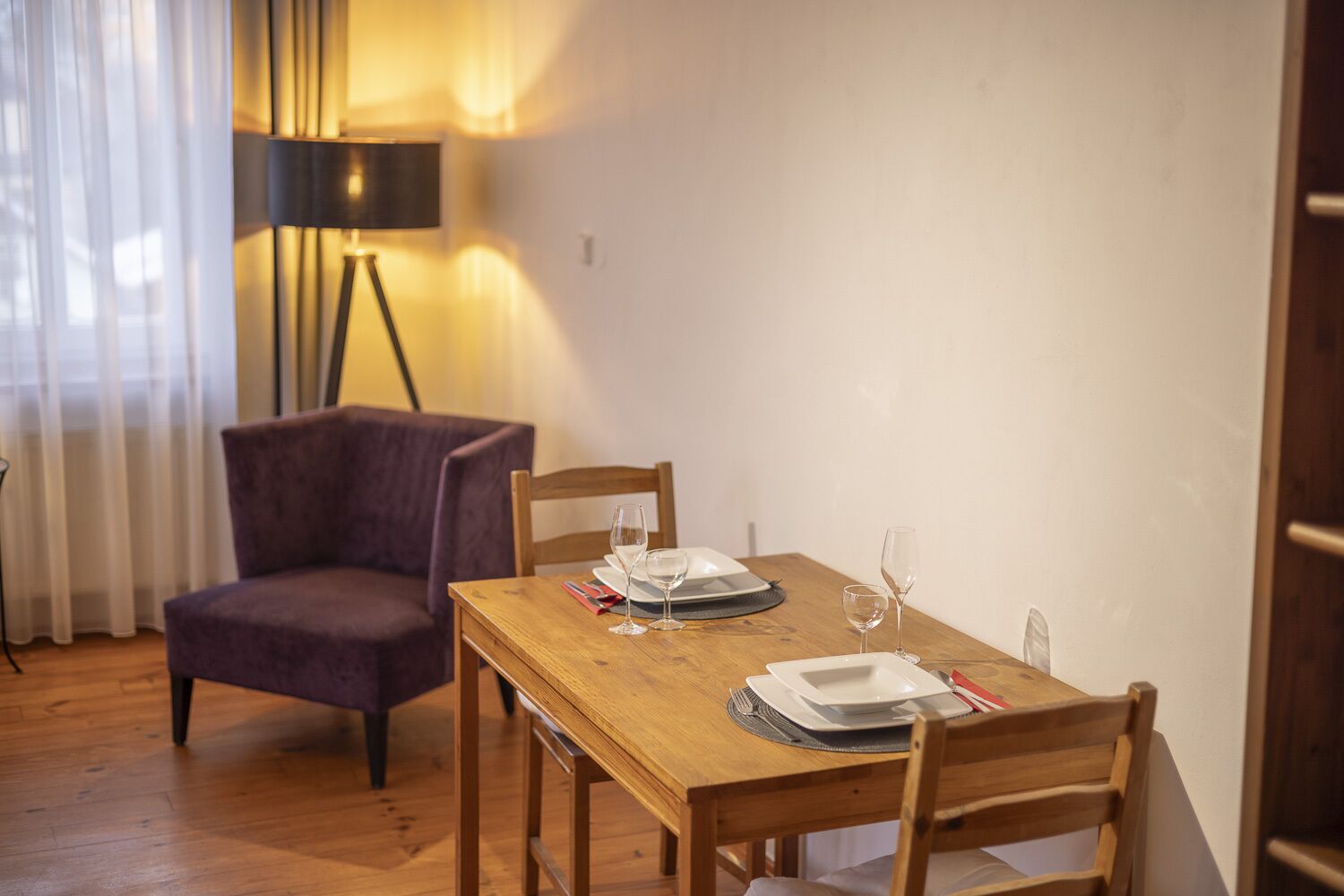 Un comedor con una mesa de madera dispuesta para dos personas, un sillón morado y una lámpara de pie.