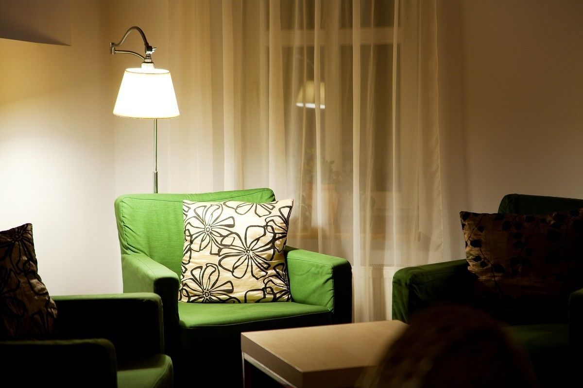 A green armchair with a patterned pillow, a small white table, and a floor lamp in a room with sheer curtains.
