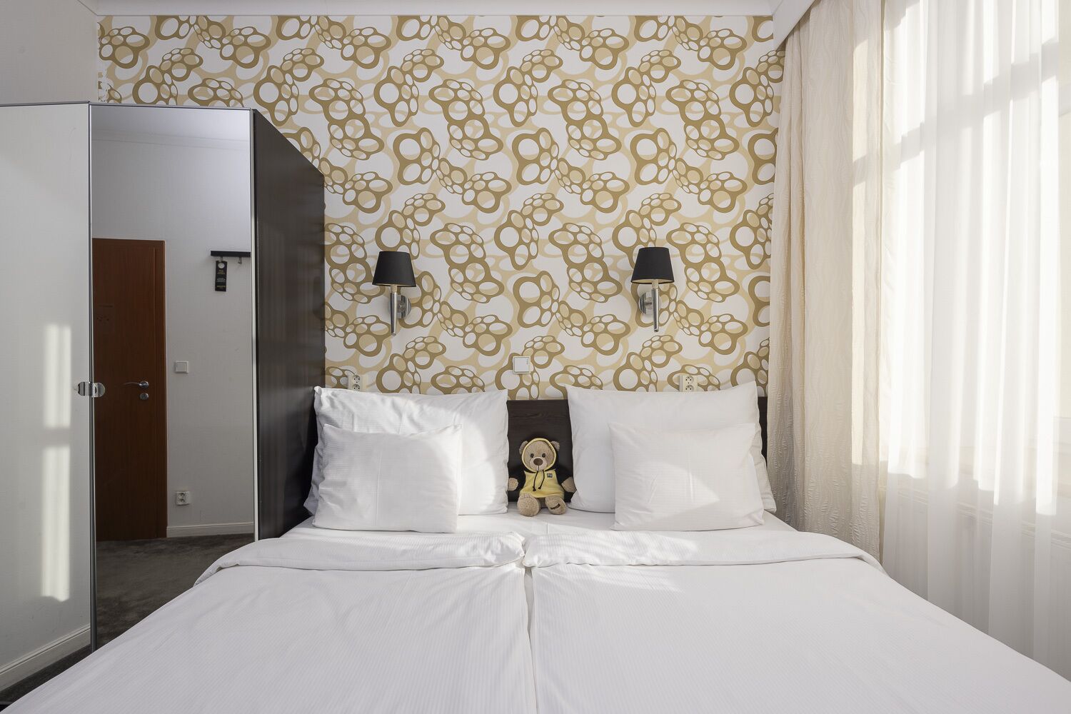 Una habitación de hotel con una cama, dos lámparas de noche, un adorno de osito de peluche y un papel tapiz estampado.