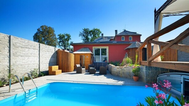 A swimming pool with a wooden deck and outdoor furniture, surrounded by a stone wall and a red building in the background.