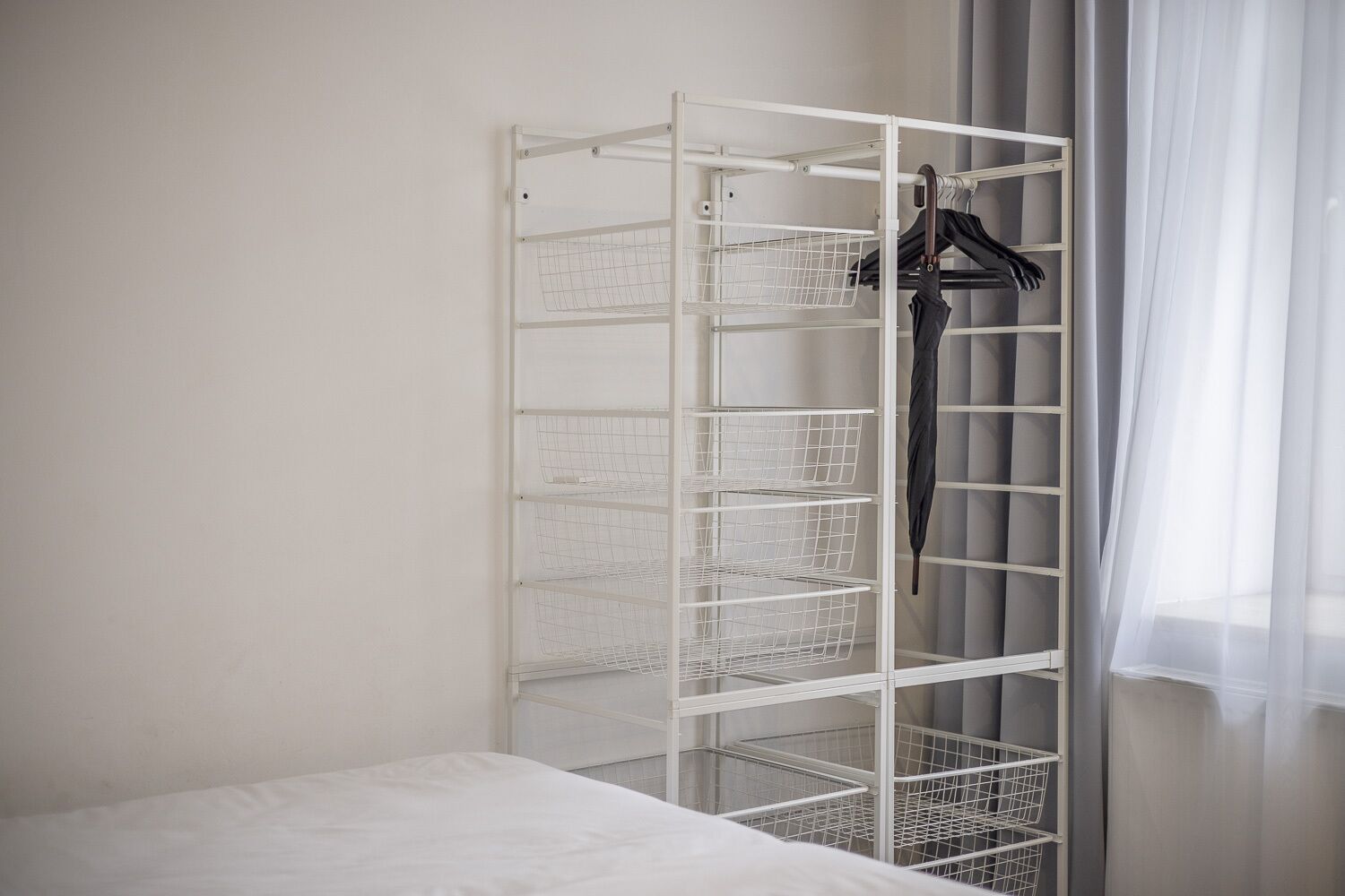 Een wit metalen rek met meerdere draadmanden, een zwarte paraplu hangt rechts, en een wit bed op de voorgrond.