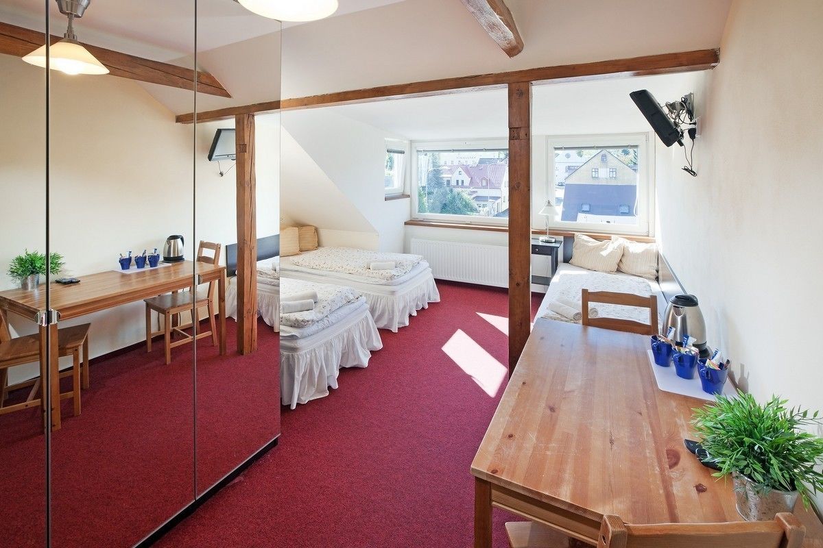 Une pièce de taille modeste, avec une table à manger en bois, deux lits blancs avec une literie assortie, une moquette rouge et une grande fenêtre offrant une vue sur les bâtiments environnants.