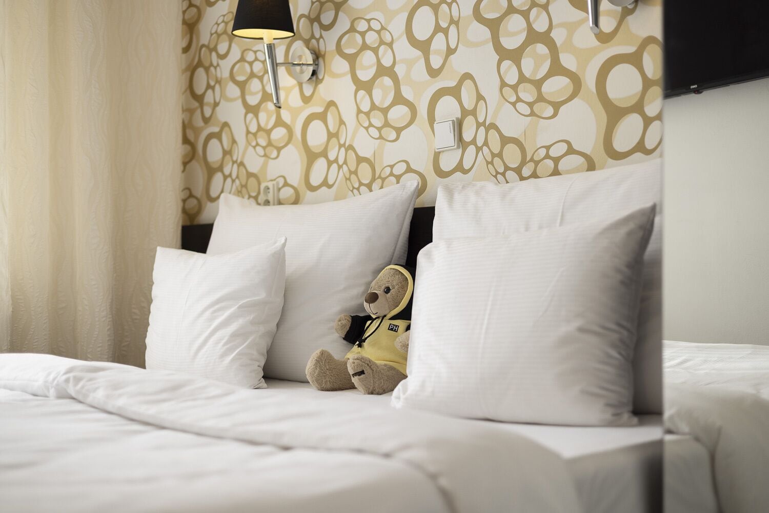 Een hotelkamer met een bed, witte kussens, een teddybeer en een wand met een rond patroon.