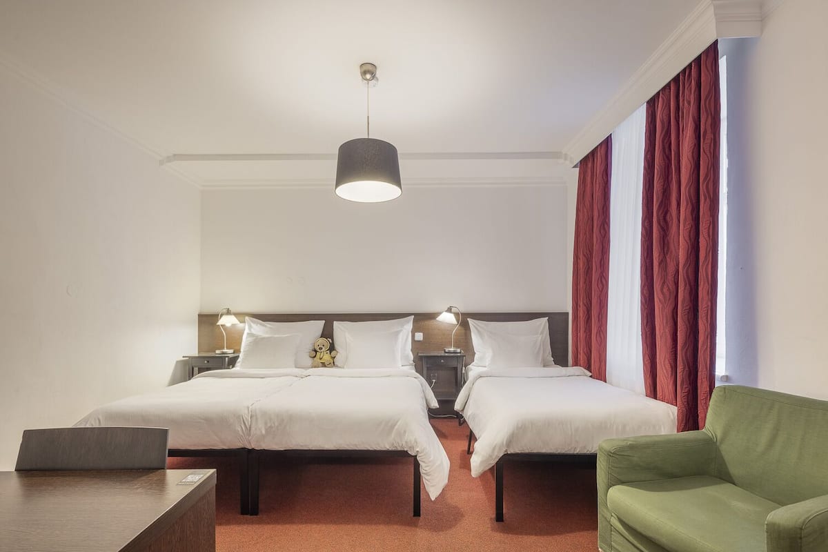 Habitación de hotel con dos camas, un escritorio, un sillón verde y un ventanal con cortinas rojas.
