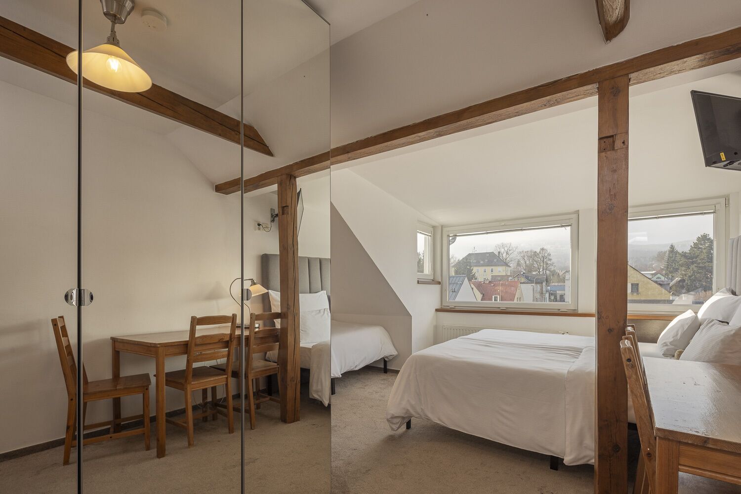 Een slaapkamer met een bed, een bureau en een stoel, een raam met uitzicht op gebouwen, en een houten balk.