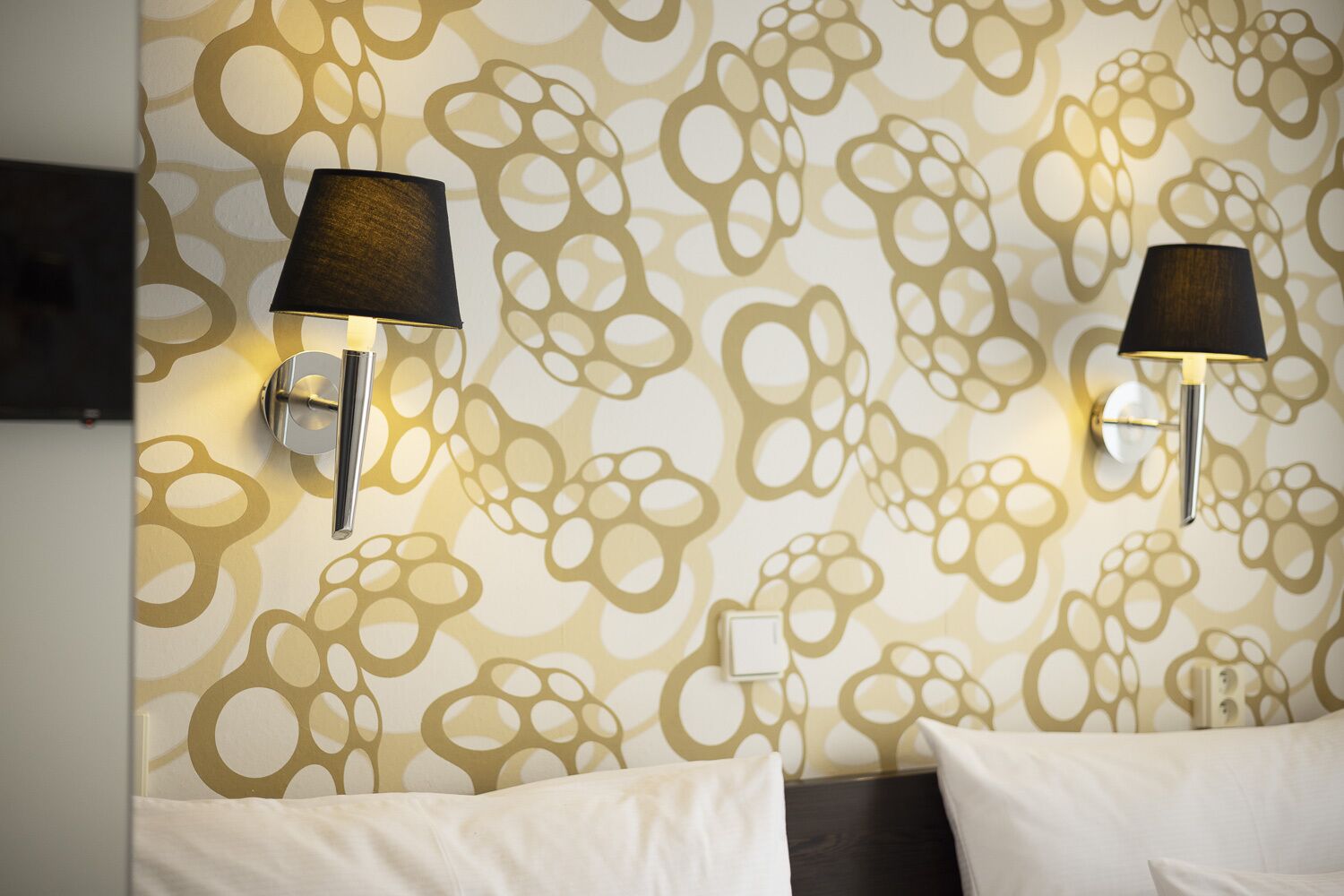 A hotel room with two wall-mounted lamps, a patterned wallpaper, and a bed with white bedding.
