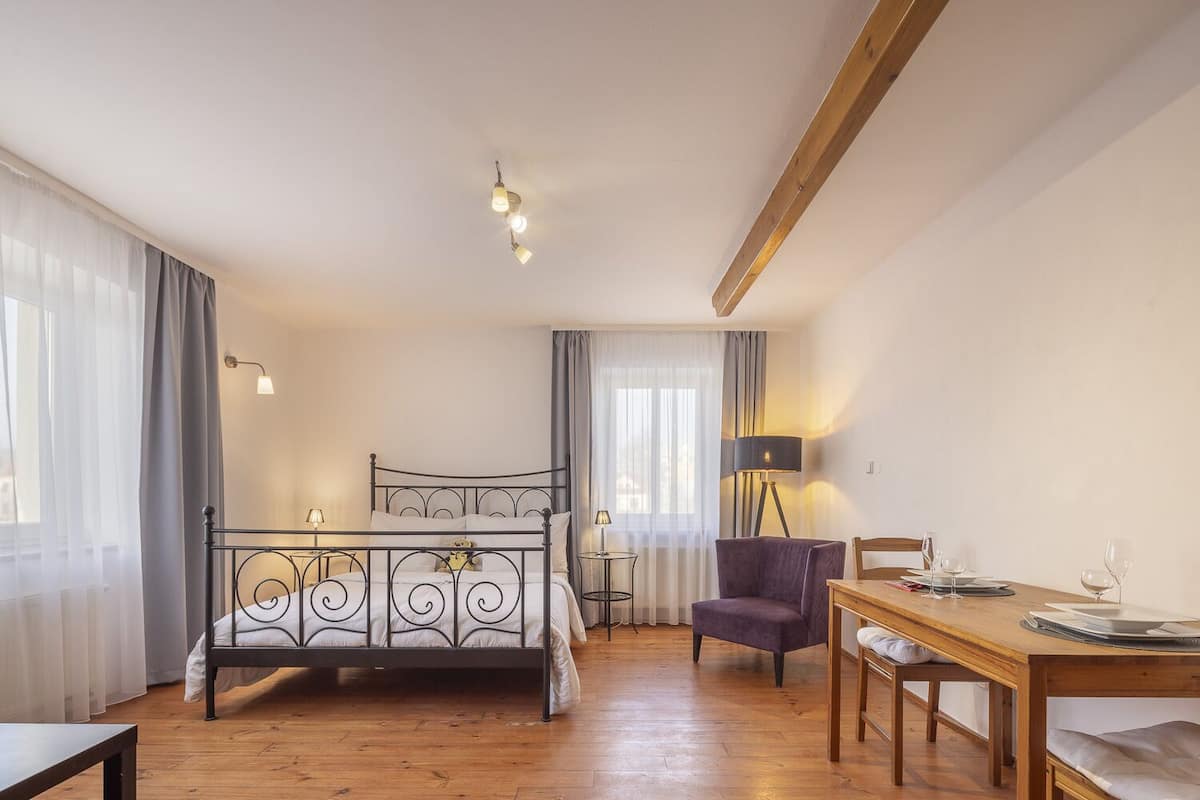 Un dormitorio con estructura de cama metálica, mesa de comedor de madera, sillón morado y una lámpara de pie.