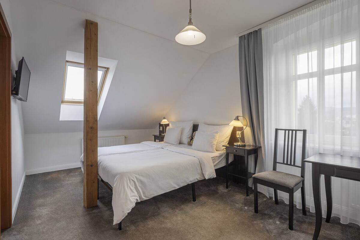 Un dormitorio con cama, mesitas de noche, una silla, escritorio, televisor, ventana y lámpara en el techo.