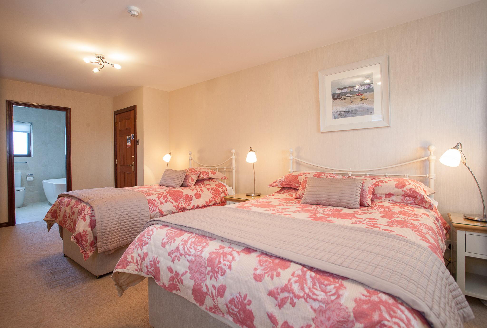 A bedroom with two beds, a bedside table with a lamp, a framed picture on the wall, and a door leading to a bathroom.
