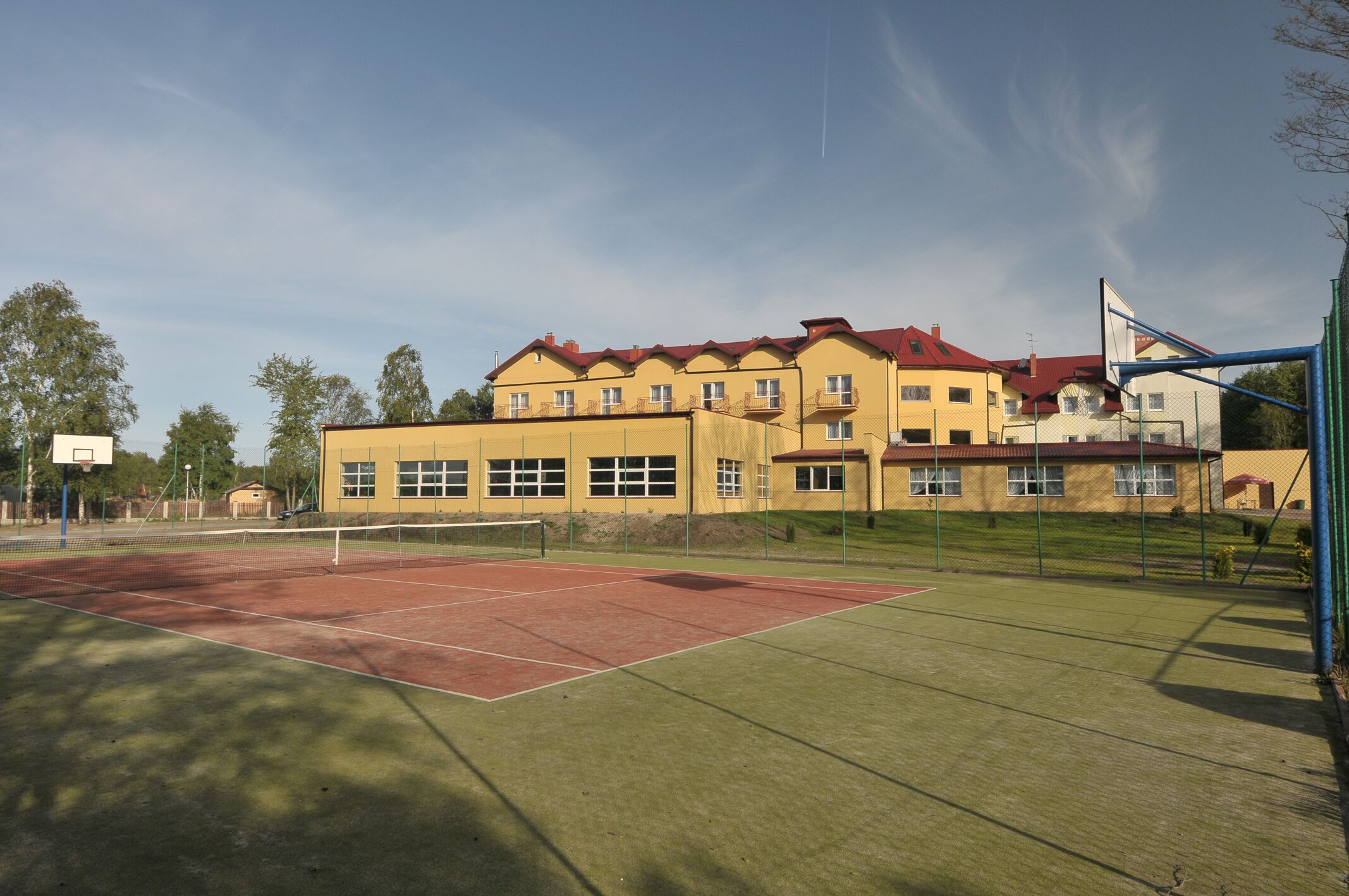 A tennis court with a net, surrounded by a fence, and a building with multiple windows and a red roof in the background.