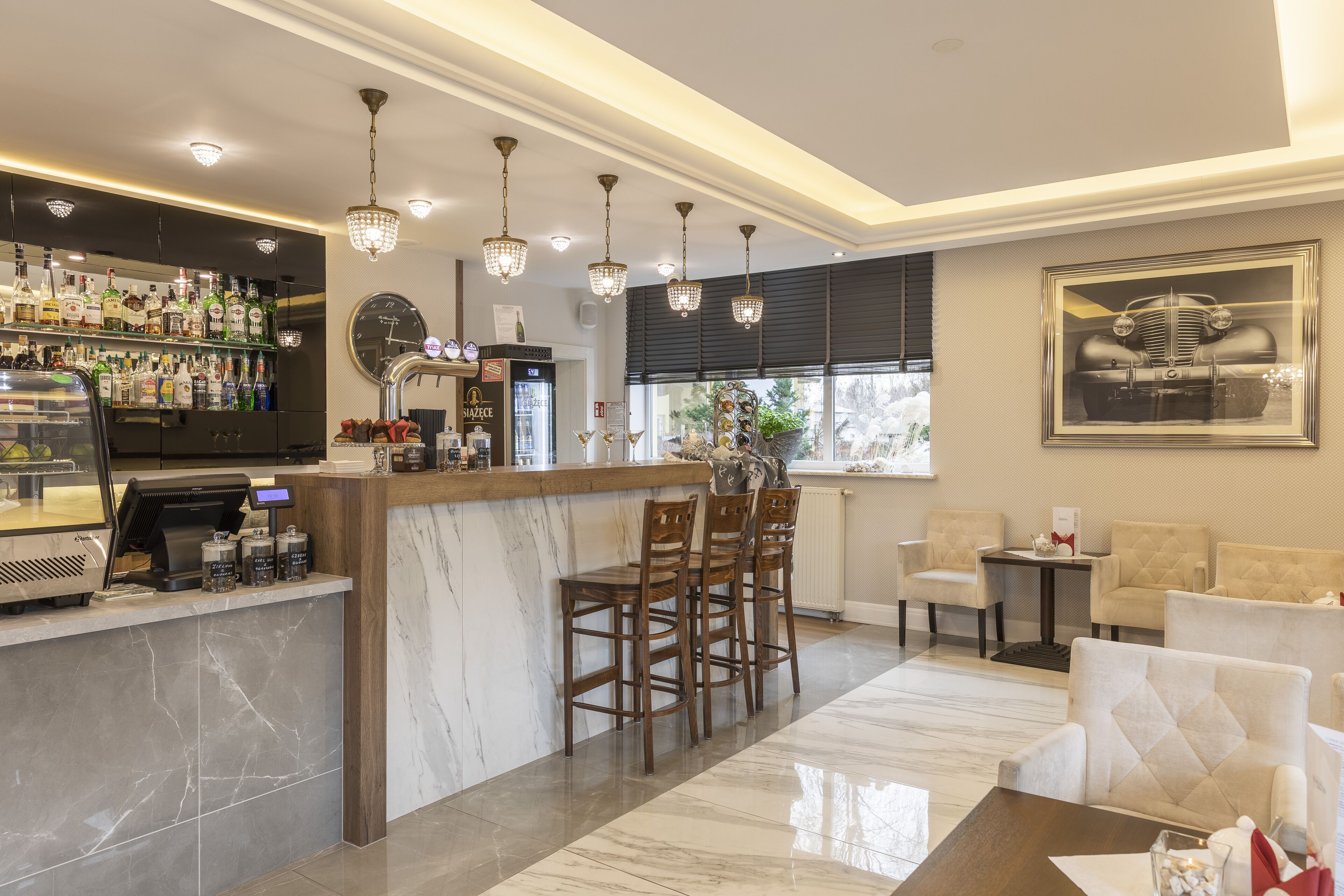 A bar area with a marble countertop, bar stools, and a variety of liquor bottles. There are hanging pendant lights and a large framed picture on the wall.
