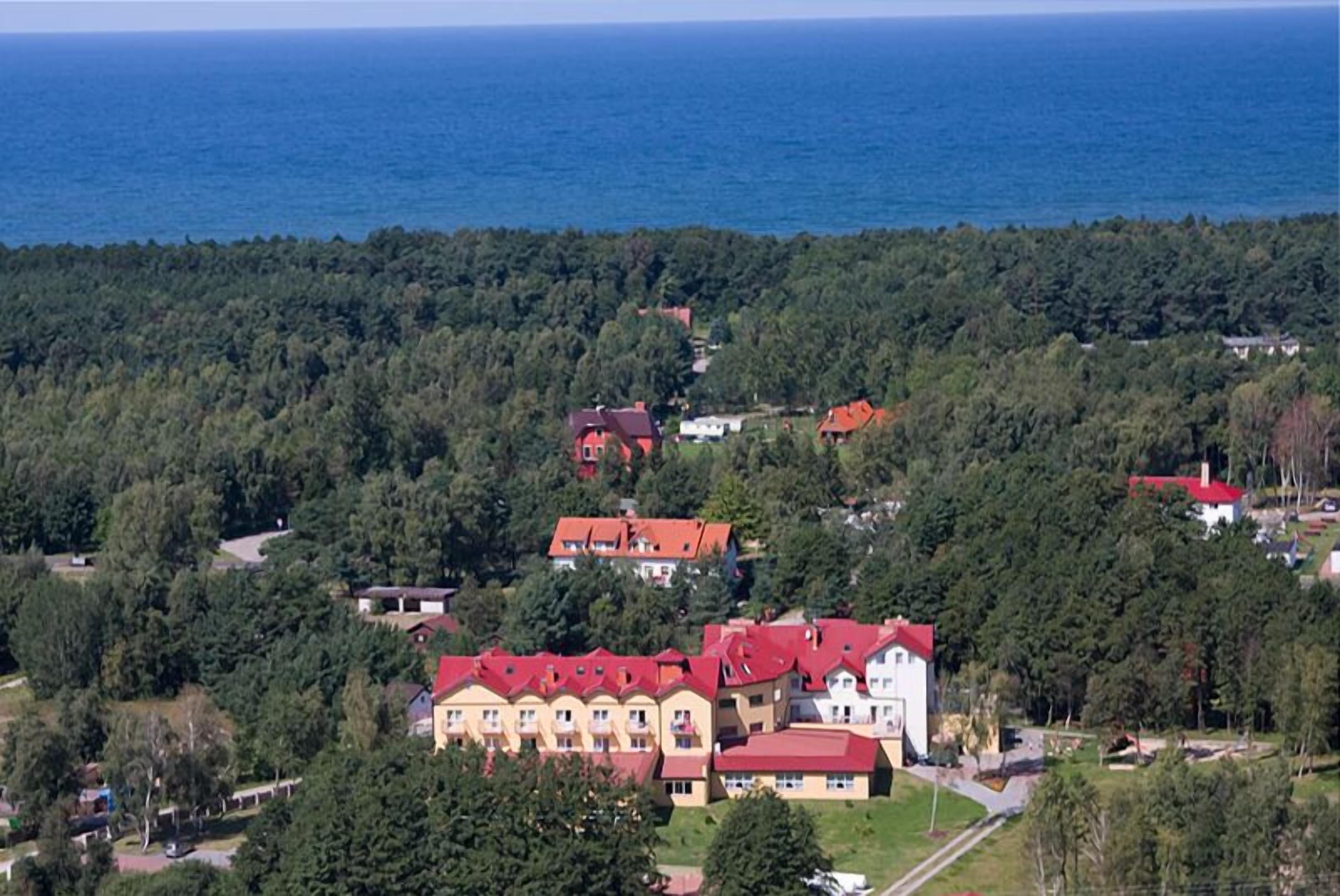 Ein großes Gebäude mit mehreren roten Dächern, umgeben von dichtem Wald, mit Blick auf ein Gewässer.