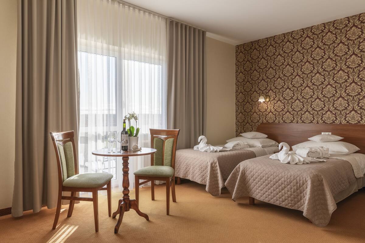A hotel room with two beds, a round table with chairs, a vase with flowers, and a patterned wallpaper.