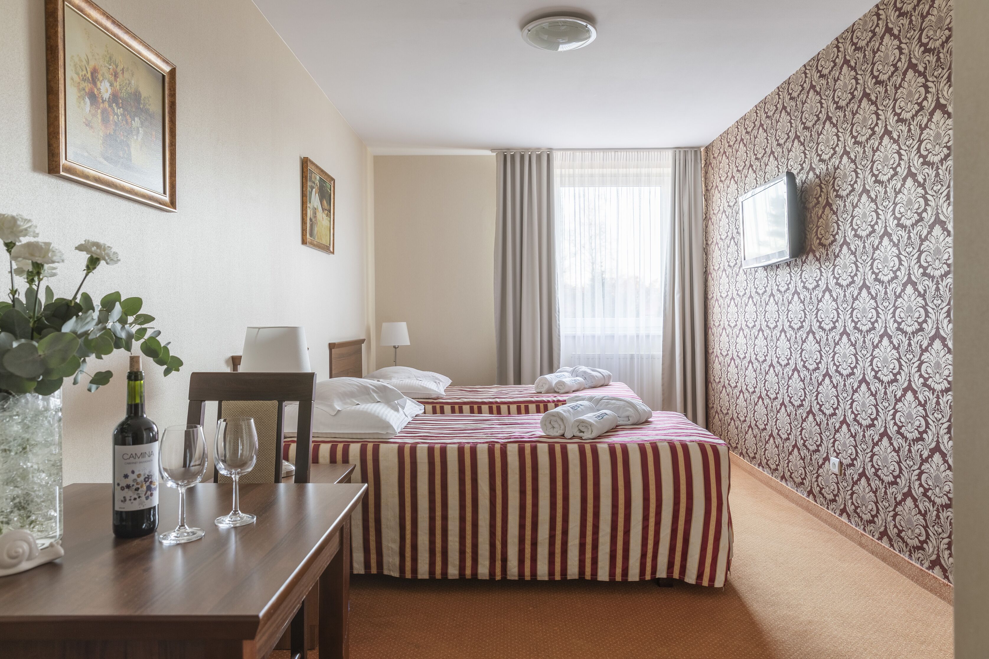 Hotelkamer met twee bedden, een televisie, een bureau met een vaas en wijnglazen, en behang met een patroon.