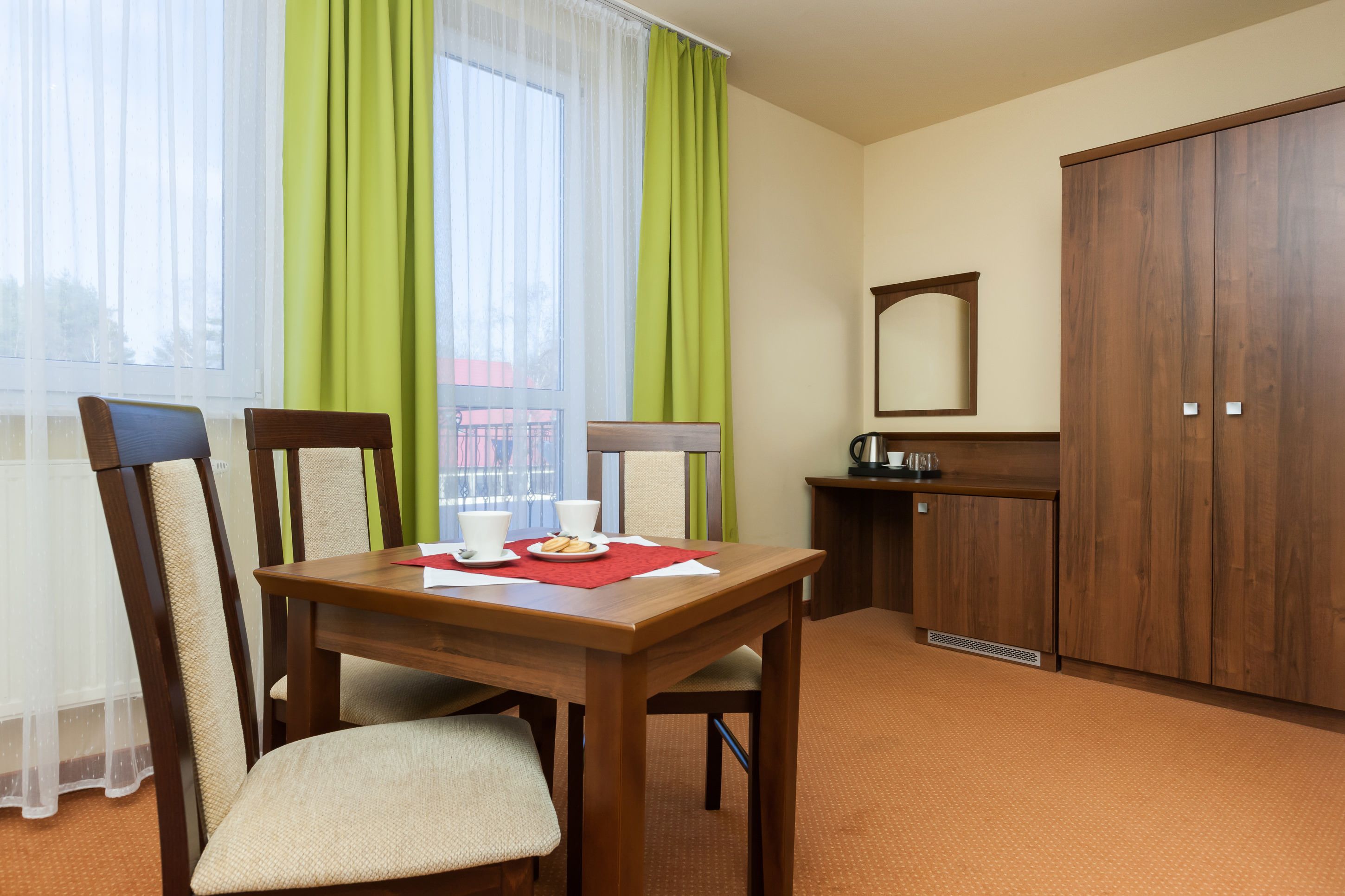 A hotel room with a wooden dining table, two chairs, a small kitchenette, and a wardrobe.