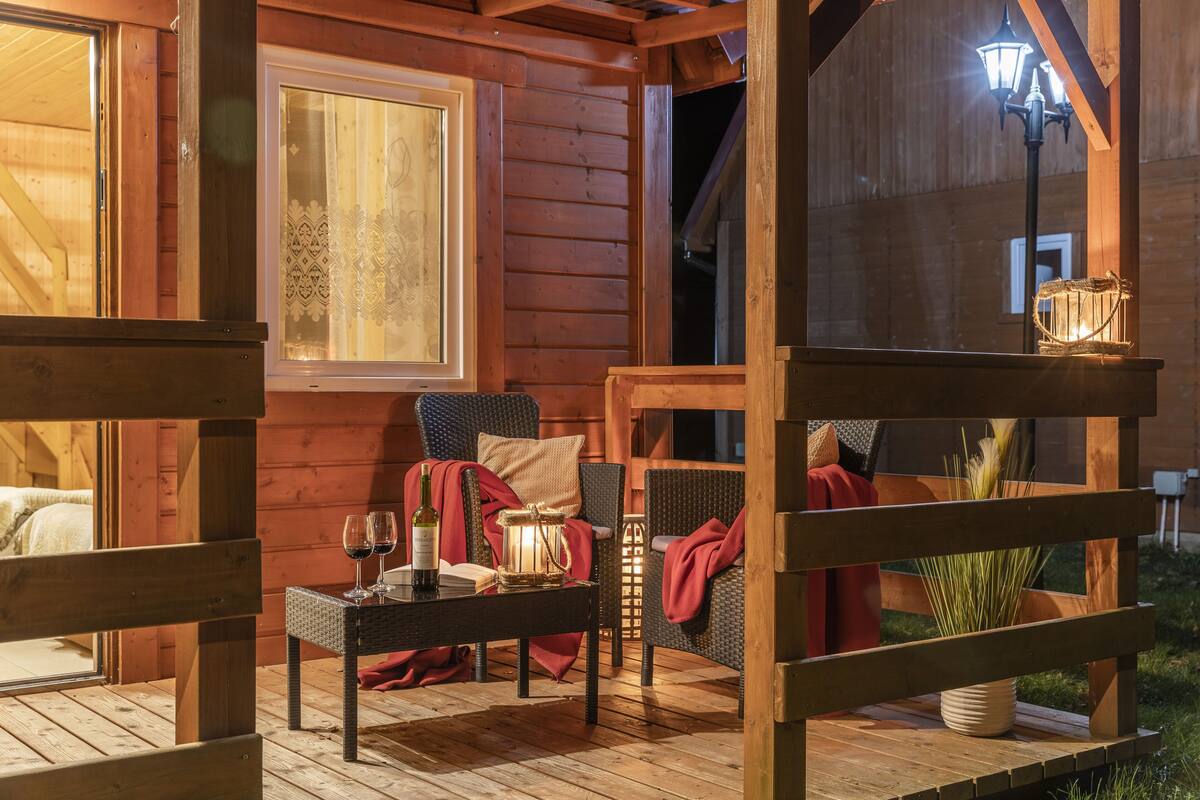 A cozy wooden cabin with a dining area featuring wicker furniture, a table with wine glasses and a candle, and a view of the surrounding forest.